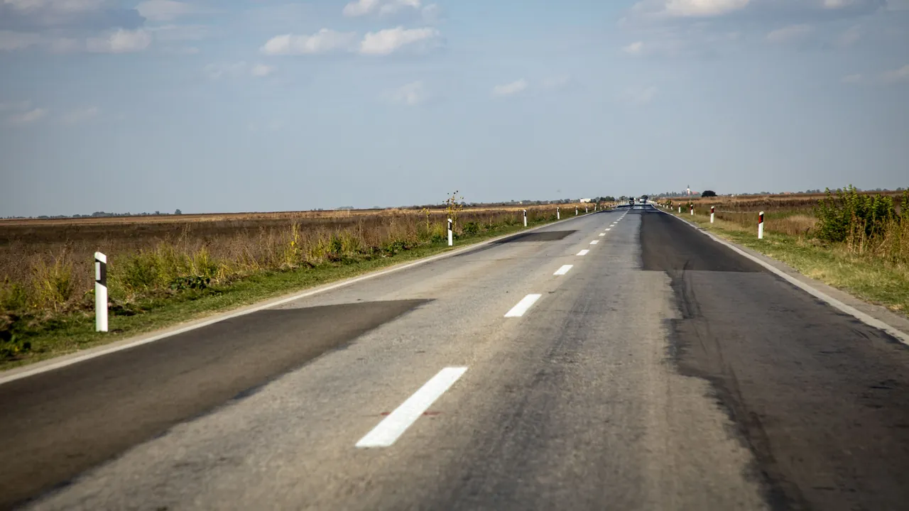 Two-lane country road with patches of darker, newer asphalt over worn gray pavement