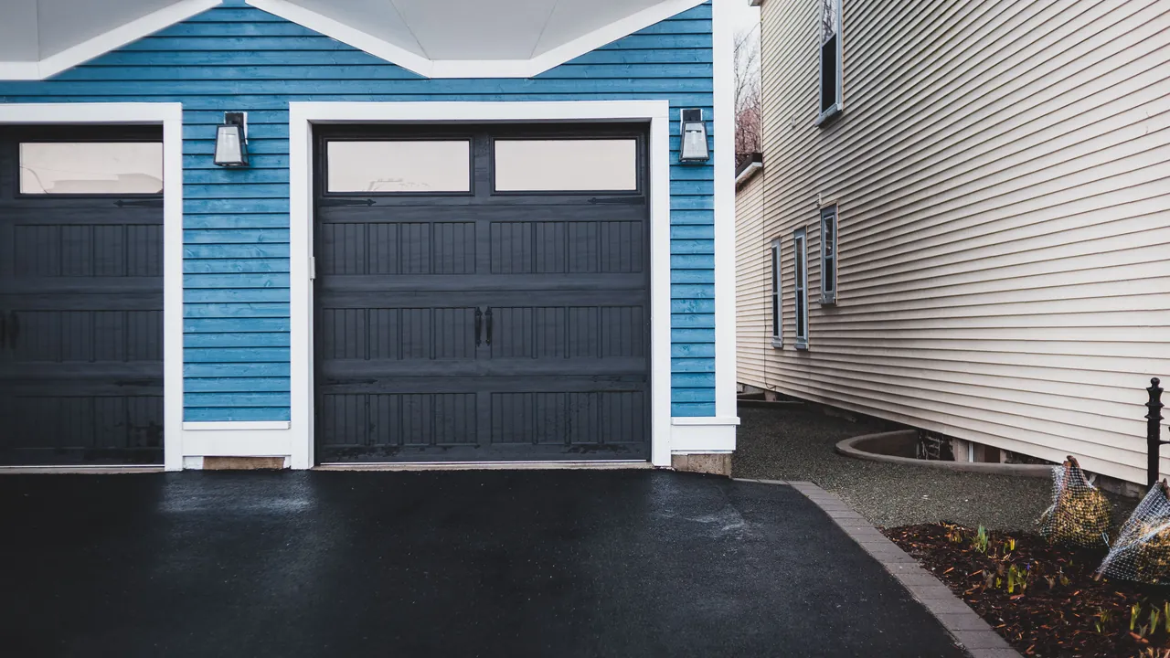 Freshly paved black asphalt driveway in front of two dark garage doors, edged with a paver border